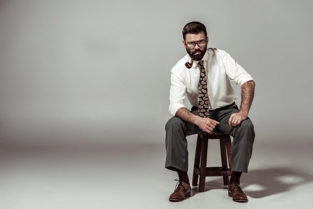 Bearded man in glasses and tie sitting on stool with pipe, highlighting premium selections at our tobacco shop in Tulsa.