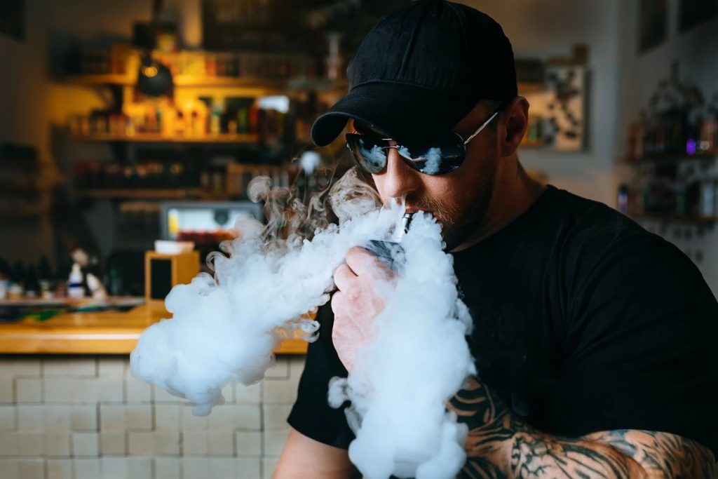 A man vaping with thick clouds in a cozy vape shop in Tulsa, surrounded by a variety of products.