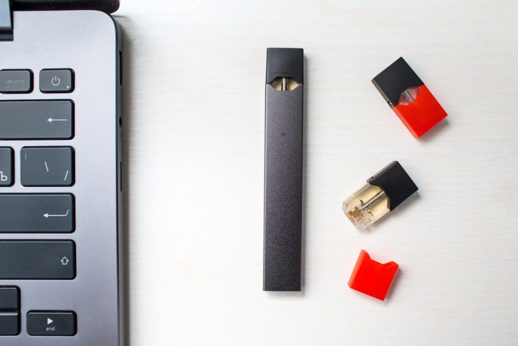 A dark gray vaporizer mod with flavored pods on a desk next to a laptop. Find the best vape mods Tulsa has to offer.