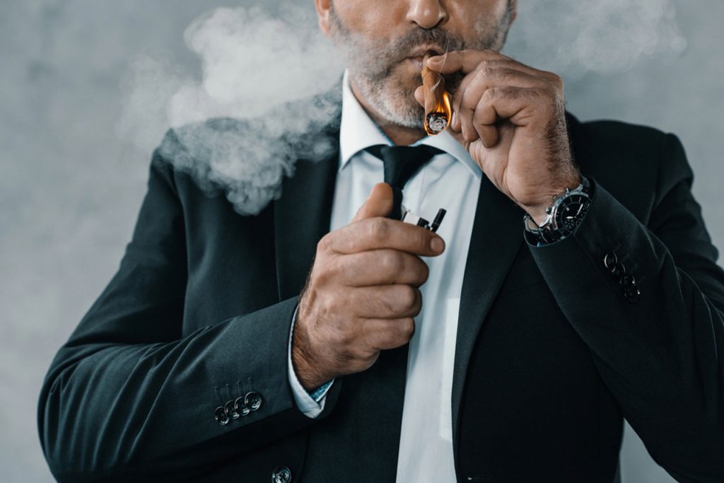 A sophisticated man in a suit lighting a luxury cigar at a cigar shop Tulsa, creating a thick cloud of aromatic smoke.
