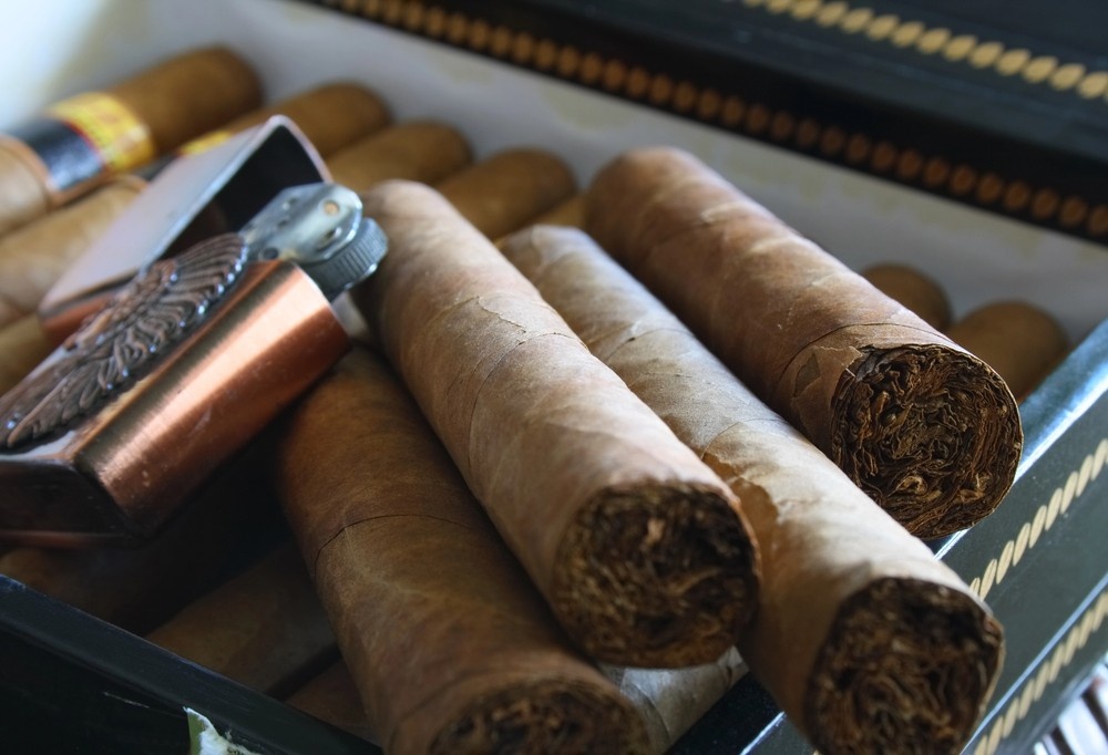 Several unbanded, hand-rolled cigars sit in a dark box alongside a copper lighter, ready for customers at a cigar shop tulsa.