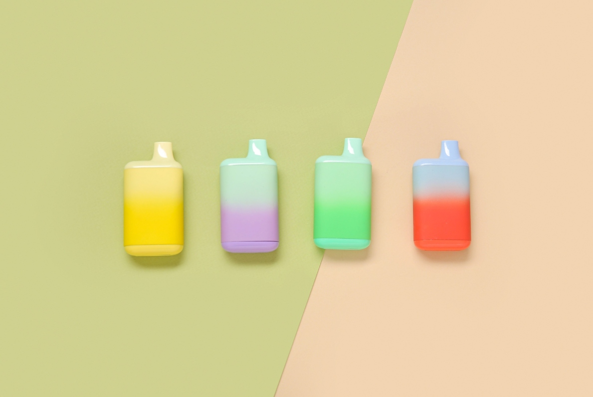 Four vibrant gradient disposable vapes in yellow, purple-mint, green-teal, and red-blue lined up on a split green-and-beige background – SMACK BOYZ bold flavors now at Tulsa's premier vape shop.