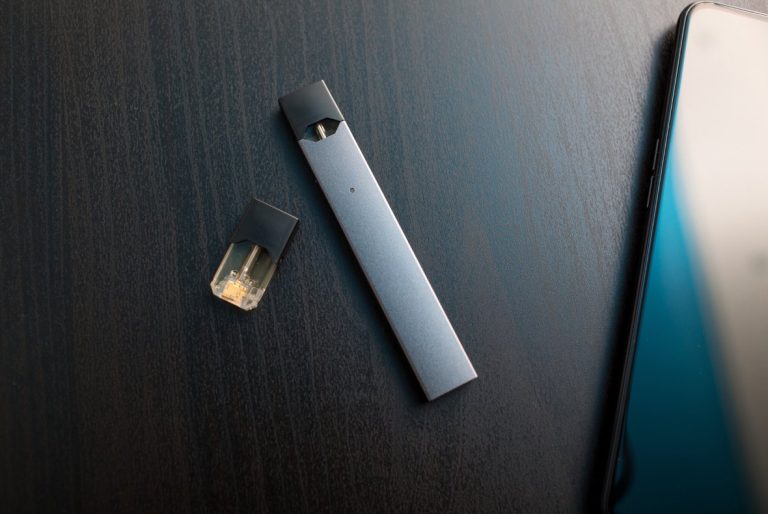 A metallic gray pod vape device and an amber e-liquid pod on a dark surface next to a smartphone. Compare vape mods Tulsa reviews.