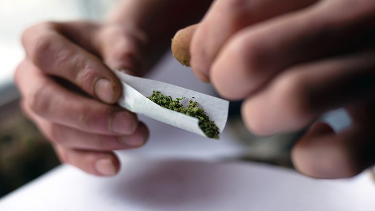 Close-up of hands carefully rolling a white cigarette paper filled with green herbs. Get premium rolling papers Tulsa delivered.