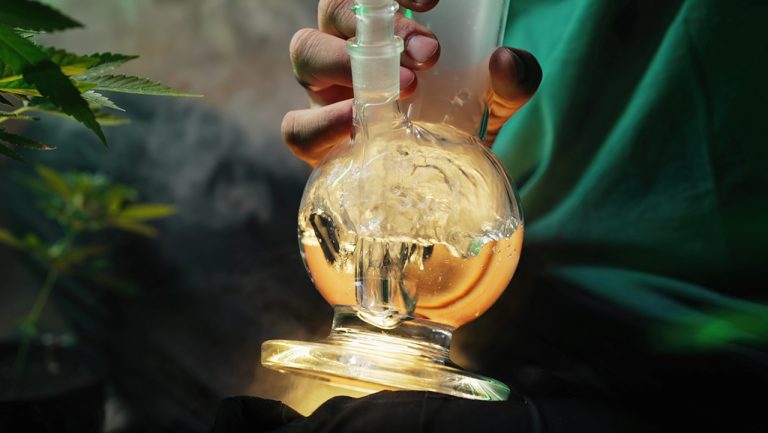 Hand holding bubbling glass pipes in Tulsa amid cannabis plants, highlighting percolator features for enhanced filtration and premium user enjoyment.